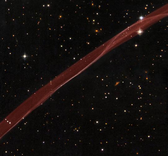 A Hubble Űrteleszkóp ACS (Advanced Camera for Surveys) műszerével a hidrogén Hα vonalában 2006 februárjában és a WFPC2 (Wide Field Planetary Camera 2) műszerével B, V és I sávban 2008 áprilisában készült felvételekből összeállított kompozit kép a szupernóva-maradvány filamentjének egy részéről (vörös sáv). A sávon belül a fényes, vékony vonal azokat a részeket jelzi, ahol a lökéshullámra pontosan a terjedési irányra merőlegesen látunk rá. Az optikai tartományban a maradvány csak a Hα vonalra érzékenyített szűrővel detektálható. A képre komponált, csillagokat és galaxisokat egyaránt tartalmazó háttér a WFPC2 kamera fenti három hullámhossz-tartományban készített képeiből állt össze. [NASA, ESA, Hubble Heritage Team (STScI/AURA)]