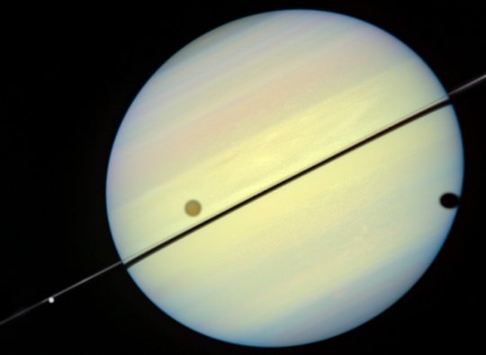 Az éléről látszó gyűrű az 1995-ös átfordulás idején. A bolygó előtt látható mustársága színű hold a Titan, melynek árnyéka a bolygókorong szélére vetül. A bal oldalon a gyűrűhöz tapadó fehér színű hold a Tethys (NASA/ESA)